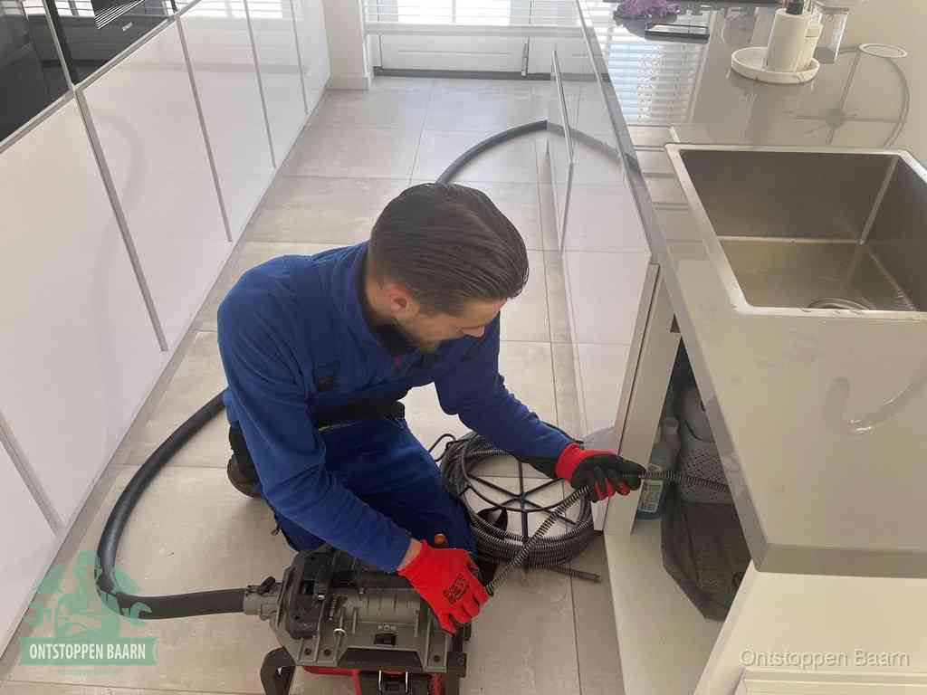 Een monteur ontstopt de keukenafvoer onder de spoelbak met een elektrische veermachine in Baarn