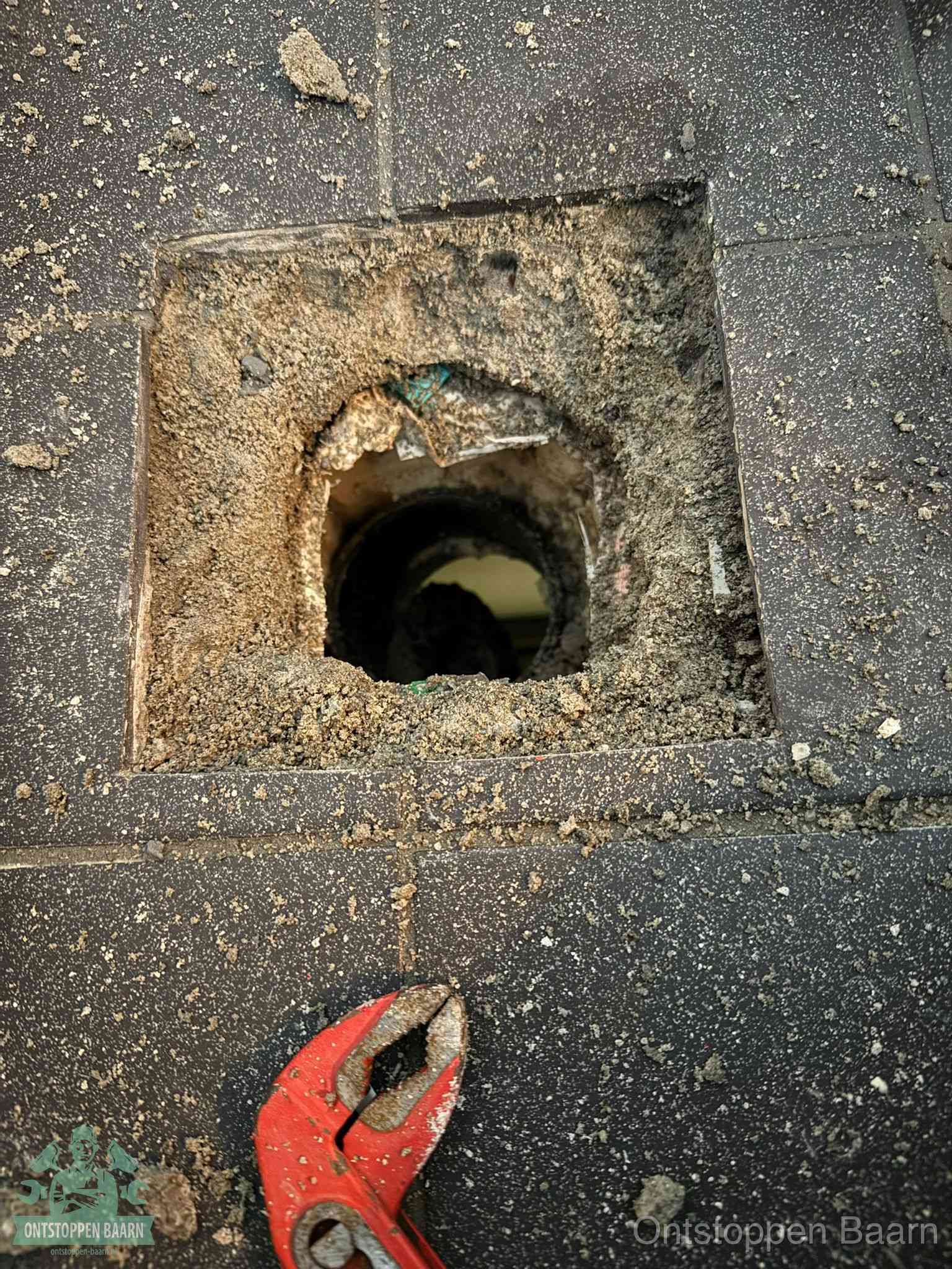 Een geopend rioolputje in een betontegelvloer met losgemaakt rooster en een zichtbare ronde afvoerbuis, het achterhalen waar de afvoerverstopping zit in Baarn