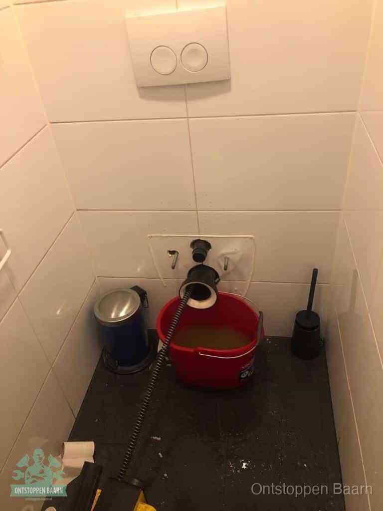 Een ontstopte wand-wc met verwijderde toiletpot waar een ontstoppingsveer in de wanduitgang verdwijnt, het achterhalen waar de verstopping zit in Baarn
