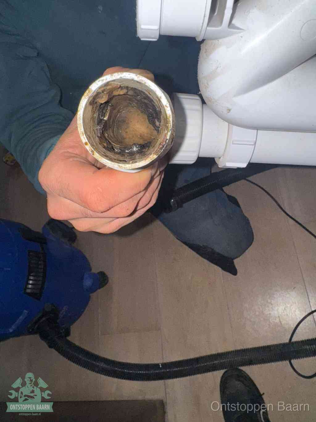 Een hand houdt een losgekoppeld PVC-afvoerbuisstuk vast dat van binnen volledig is dichtgeslibd met vet, vuil en aanslag, de meest voorkomende oorzaak van een verstopte afvoer in Baarn