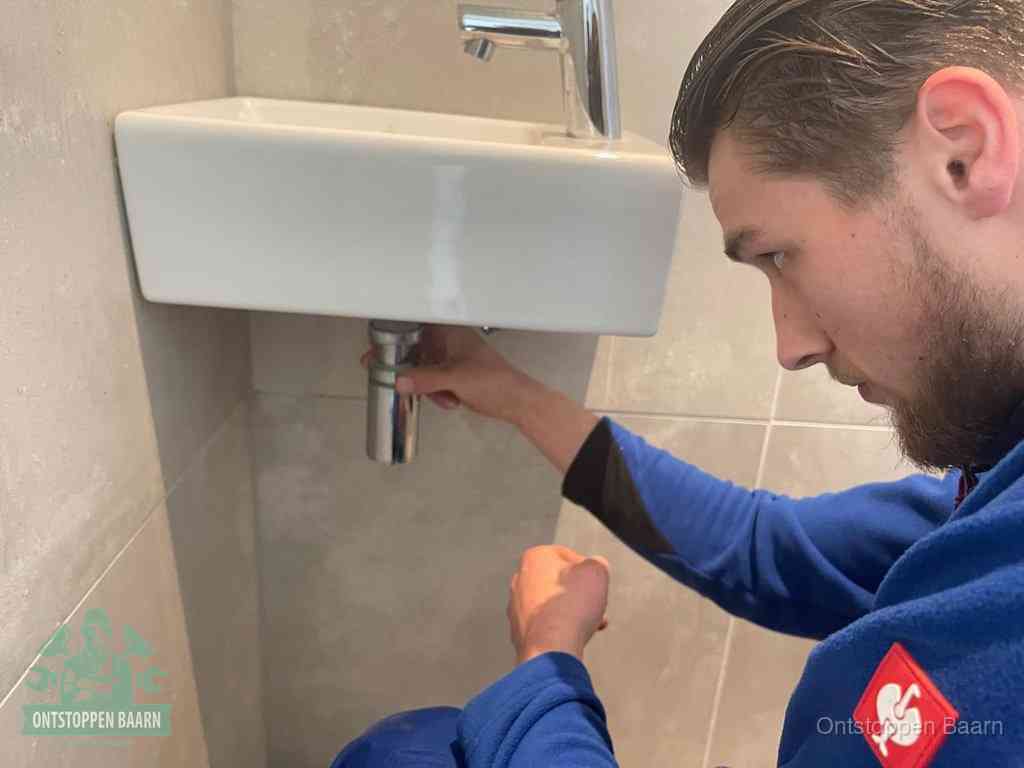 Een monteur draait met de hand de zwanenhalssifon onder een fonteinwasbak in de toiletruimte los om de bron van rioollucht in Baarn te vinden