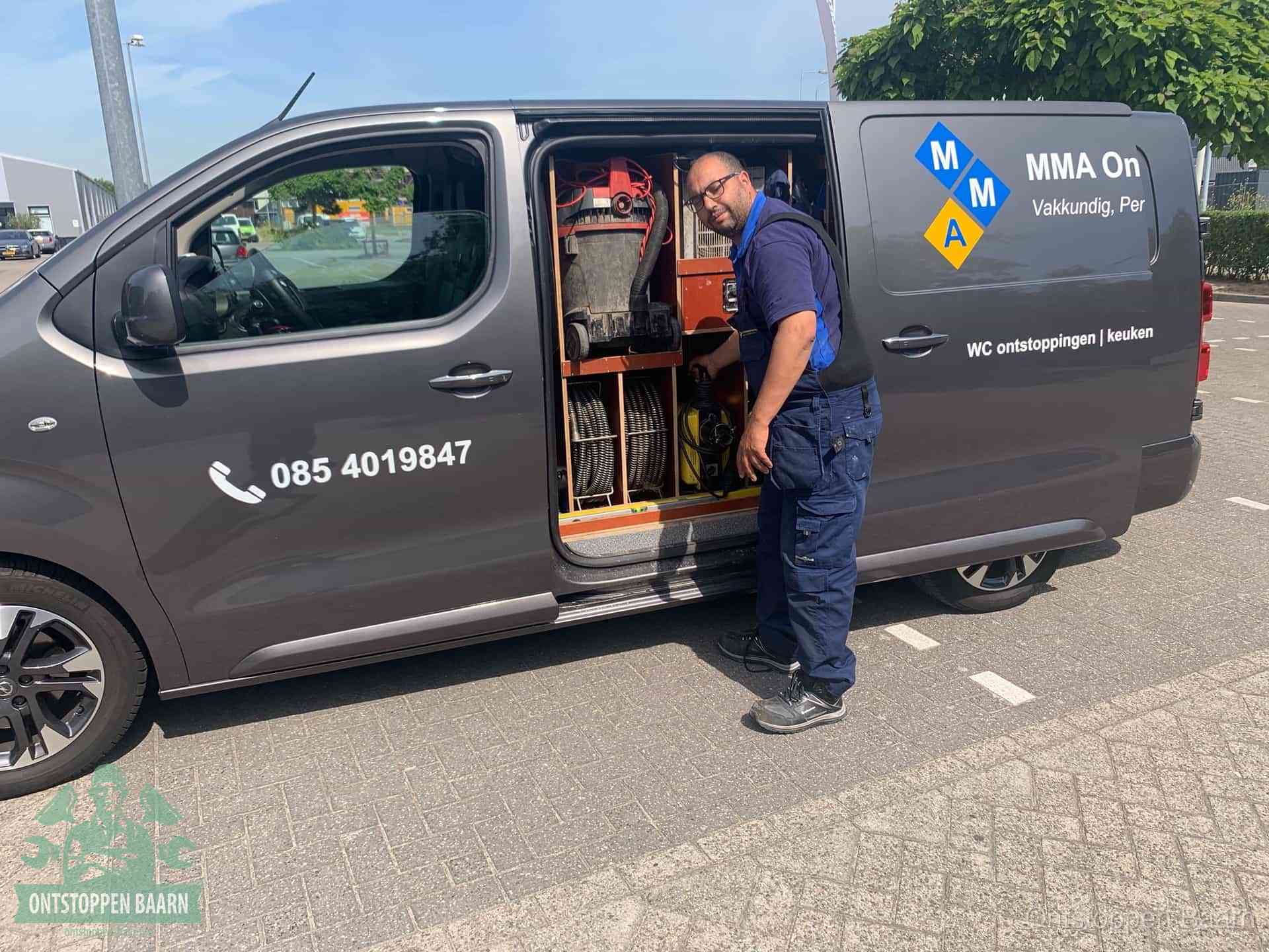 Een monteur bij de open schuifdeur van zijn bedrijfsbus op een parkeerplaats, klaar om de oorzaak van terugkerende afvoerverstoppingen aan te pakken in Baarn