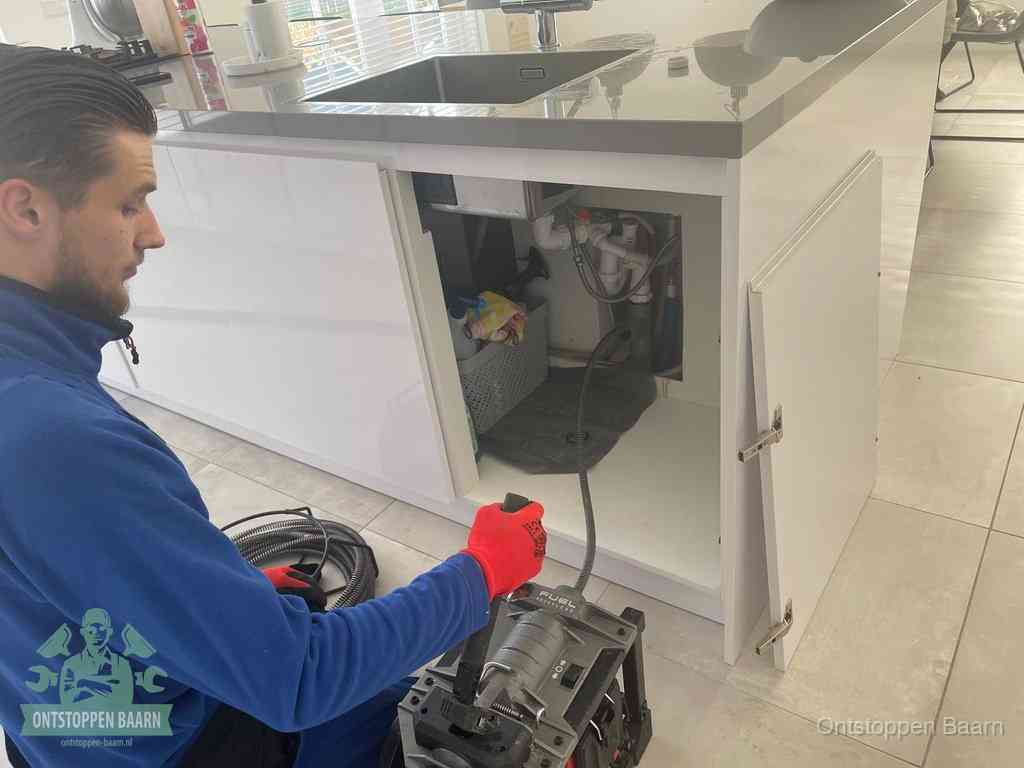 Monteur ontstopt met een veermachine een verstopte afvoer onder de gootsteen in Baarn