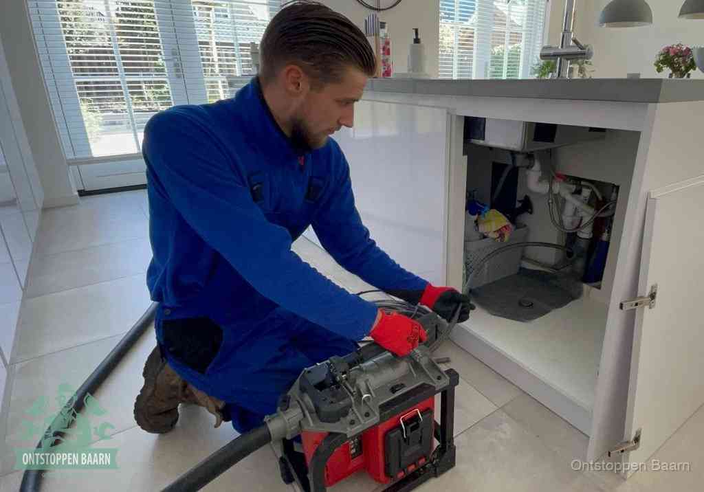 Een monteur voert een professionele elektrische ontstoppingsmachine in de afvoer onder de spoelbak tijdens het gootsteen ontstoppen in Baarn